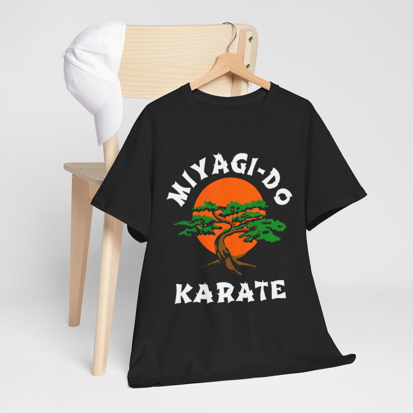 Chic display of a black Cobra Kai Miyagi-Do Karate graphic t-shirt draped over a minimalist wooden chair, accompanied by a white cap, embodying modern style.