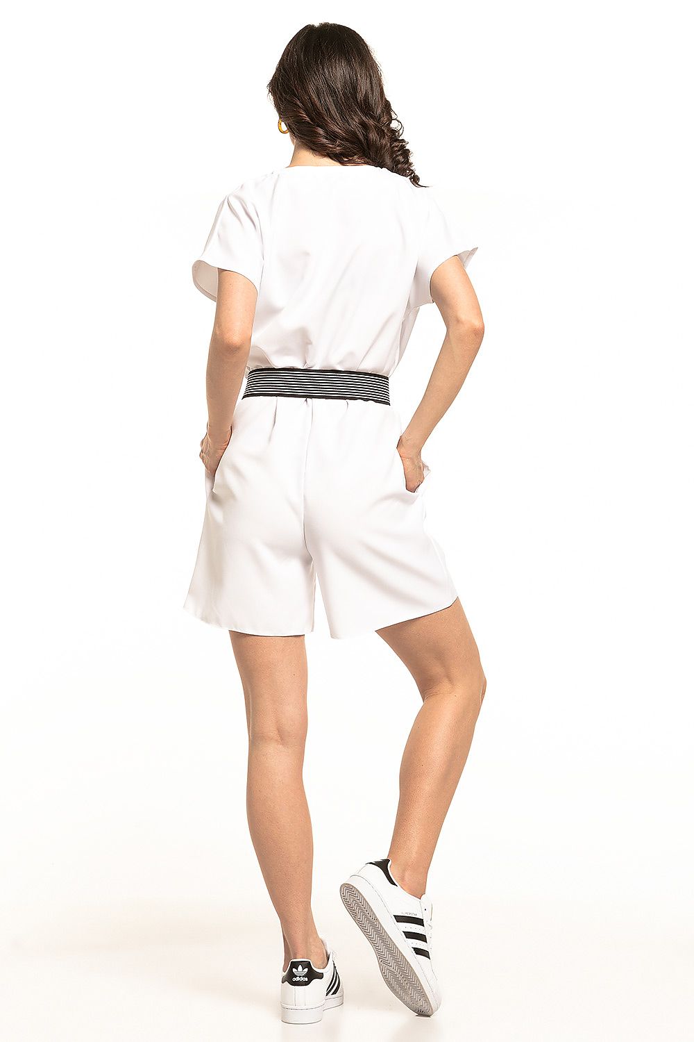 A model showcases a luxury summer jumpsuit by Tessita, featuring short sleeves and a gathered waist. The jumpsuit is styled in white with two practical pockets, perfect for warm weather, and is paired with sporty white sneakers for a chic yet casual look.
