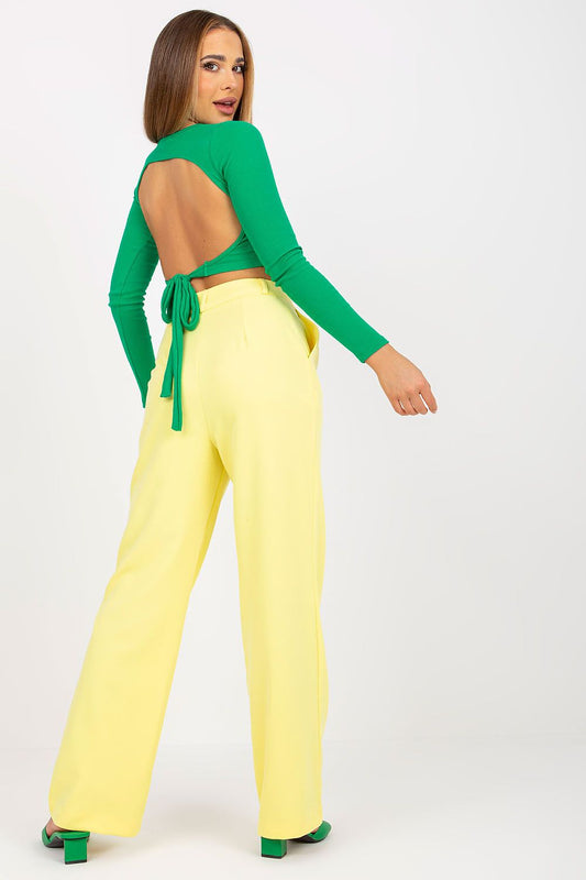 Back view of the model wearing Luxurious Wide Leg Women’s Trousers by Rue Paris, featuring a chic green top with an open back and a stylish combination with the yellow trousers, enhancing a fashionable look.