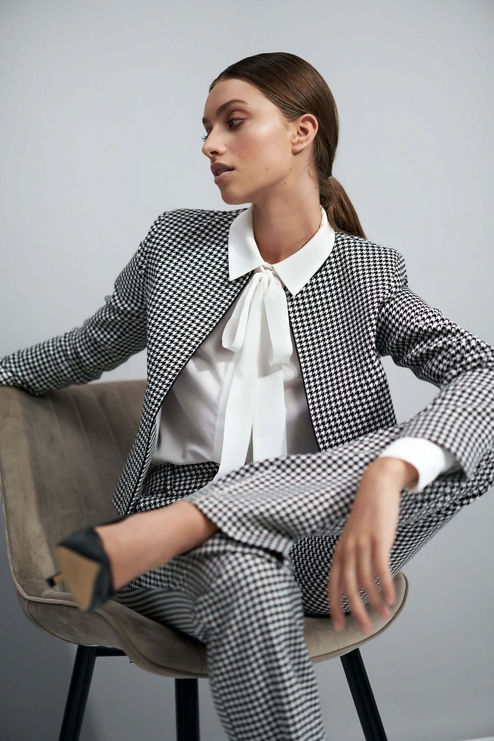 Luxury Nife Peplum Jacket for Women - Elegant Viscose Blend, Minimalist Style, Perfect Office Wear. The model is seated wearing a stylish houndstooth patterned peplum jacket paired with a white blouse and checkered trousers, exuding sophisticated elegance.