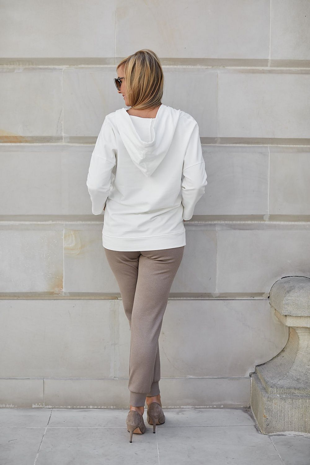 Back view of a fashionable woman in an exquisite beige hoodie, complemented by chic trousers and elegant footwear, radiating sophistication as she poses in front of a plain backdrop.