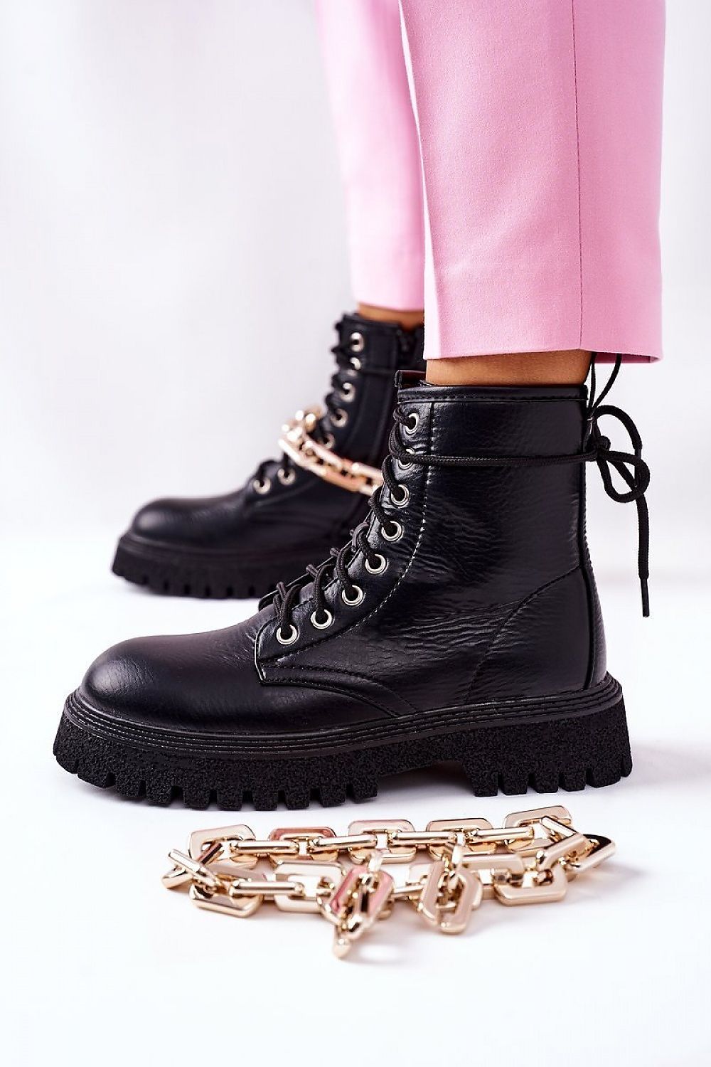 Side view of luxury women's eco-leather black boots with a bold gold chain detail, complemented by fashionable pink trousers.