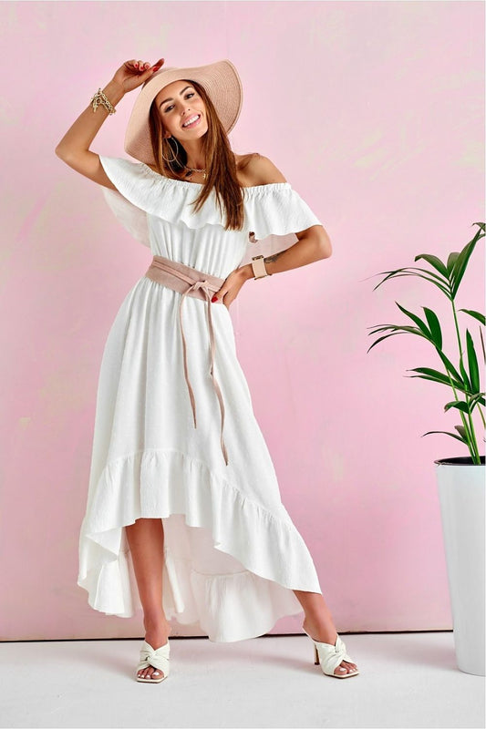 An elegant woman in a white asymmetrical maxi dress with ruffles and uncovered shoulders, styled by Roco Fashion. She poses joyfully with a wide-brimmed hat, showcasing the dress's flowy fabric and sophisticated ruffle detailing, set against a pastel pink background.