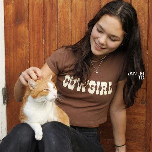 A woman sitting against a wooden backdrop, wearing a Vintage Cowgirl Print Crop Top, smiling while petting an orange and white cat, showcasing the chic summer casual style.