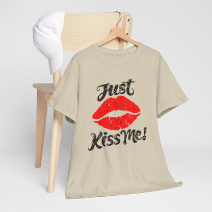 Artful presentation of a beige t-shirt adorned with a vibrant red lips graphic and the phrase 'Just Kiss Me!' staged on a minimalist wooden chair with a cap.
