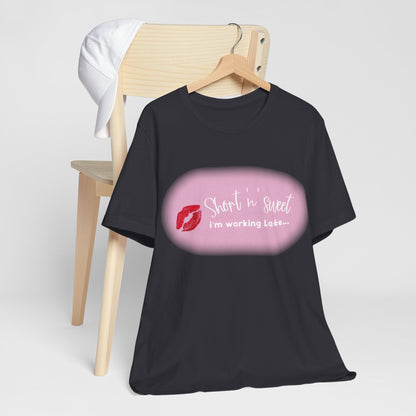 Sabrina Carpenter Inspired Unisex Short Sleeve Tee with Vibrant Lipstick Print, elegantly hung on a hanger alongside a cap, featuring the trendy phrase 'Short 'n' sweet, I'm working Late...' for a chic ensemble.