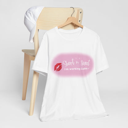Sabrina Carpenter Inspired Unisex Short Sleeve Tee, featuring a vibrant lipstick print design, elegantly displayed on a wooden hanger against a minimalistic backdrop with a white cap in the background. Perfect for stylish fans of Sabrina Carpenter.