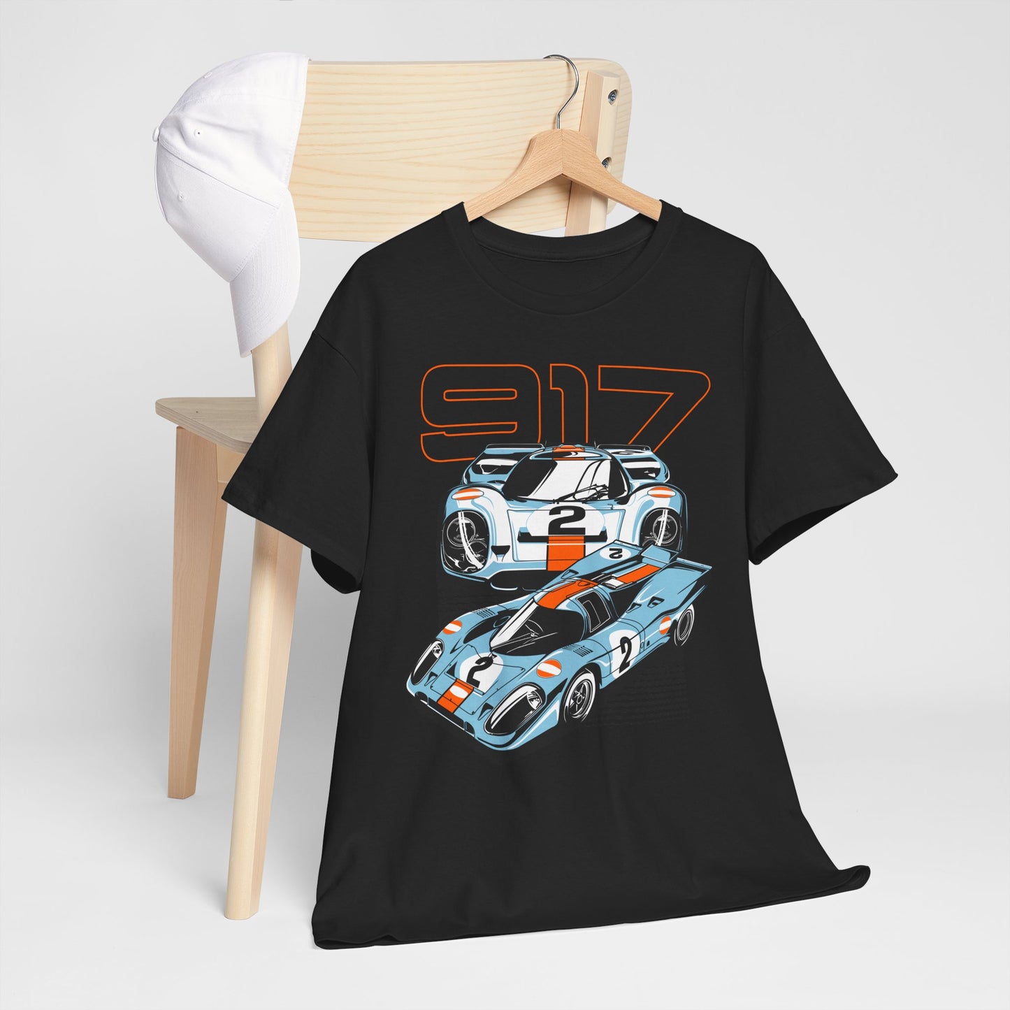Black t-shirt with Porsche 917 graphic on a wooden chair next to a white cap, presenting a stylish casual outfit for automotive aficionados.