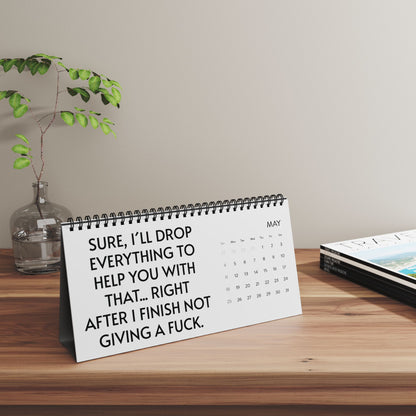 2025 Humorous Desktop Calendar for Work with Cheeky Quotes set on a wooden desk, showcasing the phrase 'SURE, I’LL DROP EVERYTHING TO HELP YOU WITH THAT... RIGHT AFTER I FINISH NOT GIVING A FUCK,' creating a fun atmosphere.