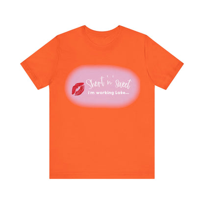 Sabrina Carpenter Inspired Unisex Short Sleeve Tee with Vibrant Lipstick Print in bright orange, featuring the phrase 'Short n' sweet i'm working Late...' decorated with a bold red lipstick mark.
