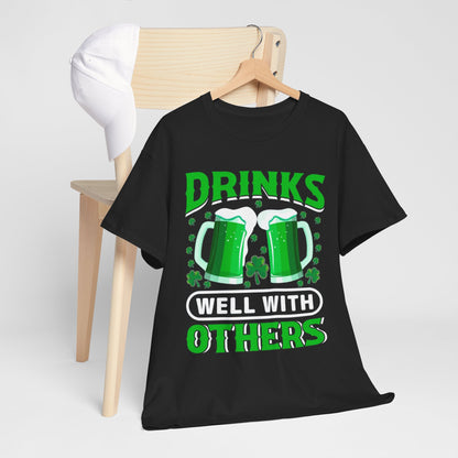 Chic presentation of a Funny St. Patrick's Day Unisex T-Shirt on a hanger with 'Drinks Well With Others' design and Irish beer mugs, exuding festive charm.