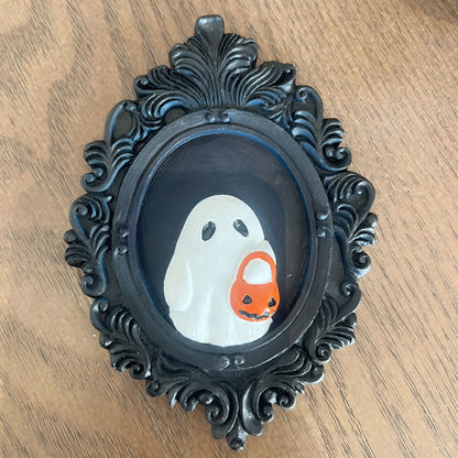 Elegant Halloween Resin Ornament - Trick or Treat Ghost Wall Decoration. A charming white ghost with a pumpkin basket adorns a decorative black frame, enhancing Halloween décor.