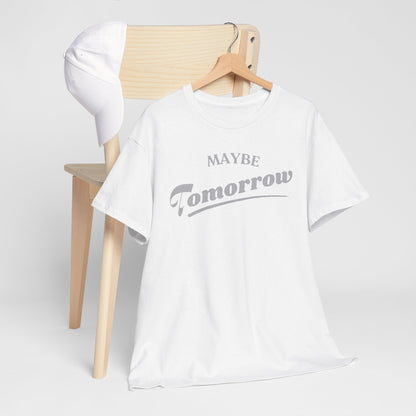 Chic presentation of a white t-shirt with the phrase 'Maybe Tomorrow' inscribed in stylish grey font, placed artistically alongside a white cap on a wooden chair.