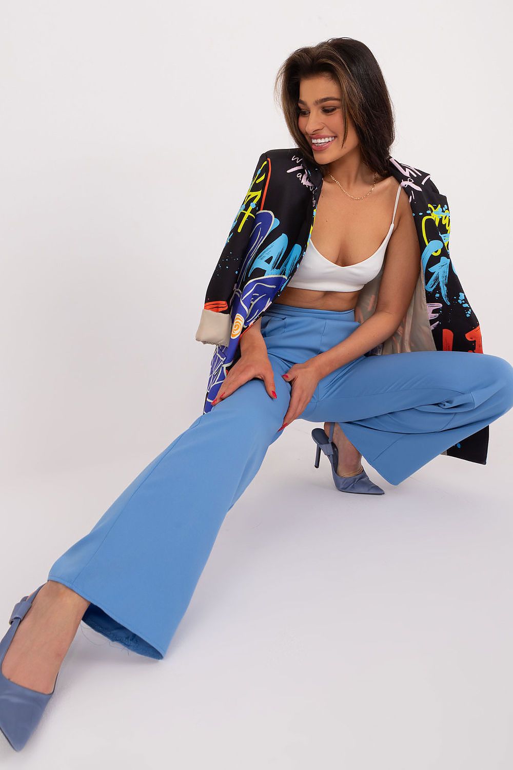 Elegant woman wearing high waist unbuttoned trousers by Rue Paris, paired with a stylish black graphic blazer and blue heels, posing confidently in a modern setting.