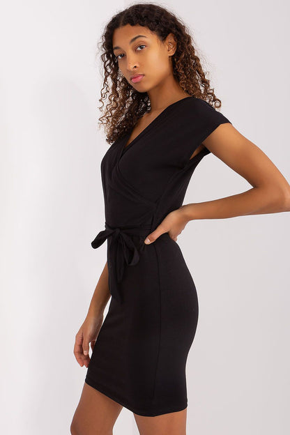 A woman stands stylishly in a black cotton summer pencil dress with a tie belt by Rue Paris, exemplifying modern elegance with her confident posture.
