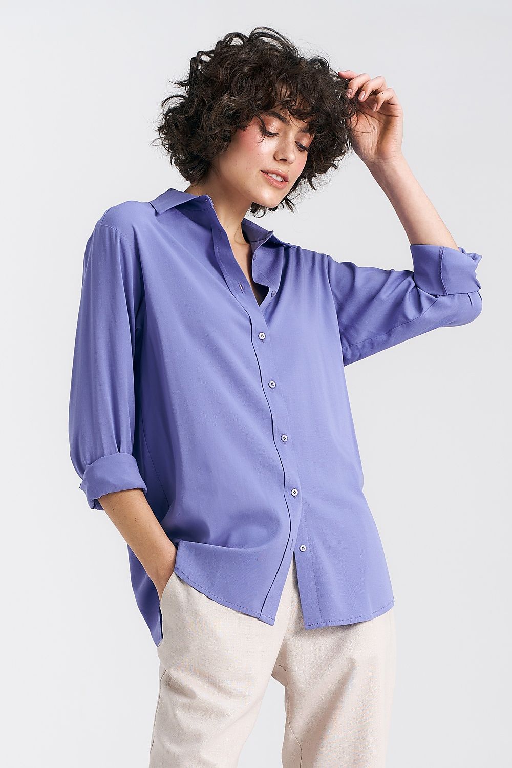 A model showcases a luxurious oversized long sleeve viscose shirt in soft lavender, featuring a classic collar and elegant button closure by Nife. The shirt is styled with casual trousers, highlighting its versatility for both sophisticated and relaxed outfits.