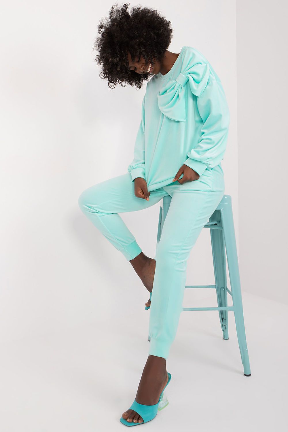 Elegant woman in the Italy Moda Velour 2-Piece Sweatshirt and Pants Set in aqua, playfully posing while displaying the stylish bow accent on the blouse.