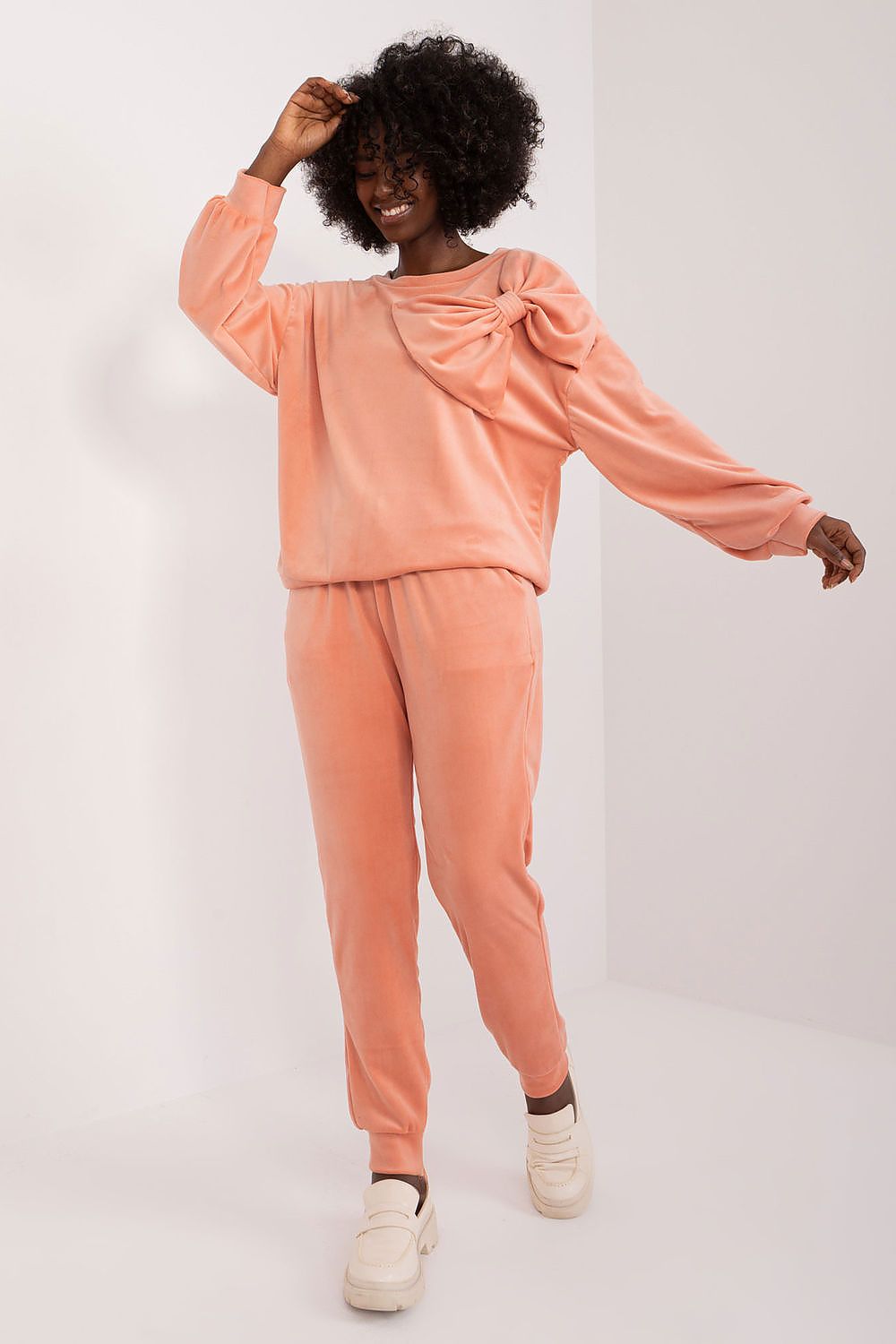 Radiant woman enjoying a light-hearted moment in the Italy Moda Velour 2-Piece Sweatshirt and Pants Set in soft peach, featuring a chic bow detail, perfect for stylish comfort.