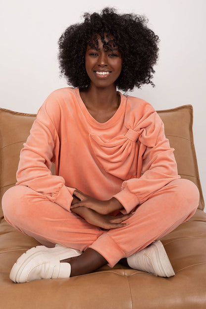 Italy Moda velour sweatshirt and pants set in peach colour, featuring a stylish bow detail on the blouse. Model exudes elegance while seated comfortably on a luxurious sofa.