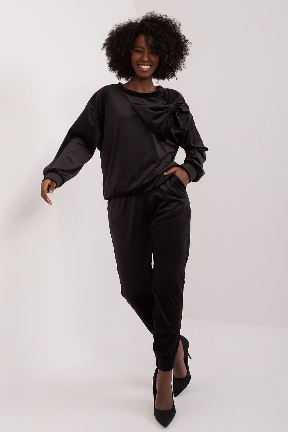 Italy Moda velour sweatshirt and pants set in classic black, adorned with a sophisticated bow detail. Model stands elegantly, embodying modern luxury with confidence in high heels.