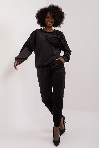 Italy Moda velour sweatshirt and pants set in classic black, adorned with a sophisticated bow detail. Model stands elegantly, embodying modern luxury with confidence in high heels.