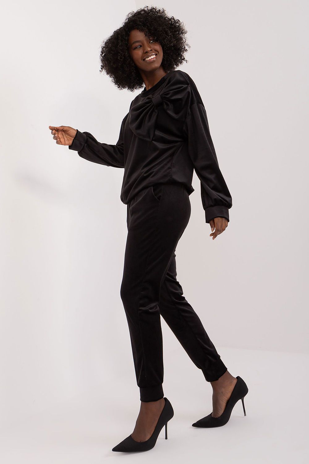 Side view of Italy Moda velour sweatshirt and pants set in black, emphasizing the stylish bow detail on the blouse. Model poses gracefully, highlighting the contemporary design.
