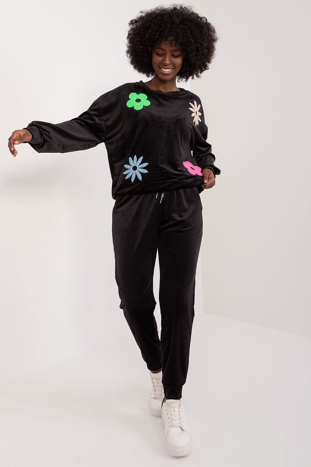 Model presenting the sophisticated black floral velour set by Italy Moda, featuring a plush long-sleeve sweatshirt with colourful floral appliqués, complemented by sleek black high-waisted pants and trendy white sneakers, exemplifying luxury loungewear.
