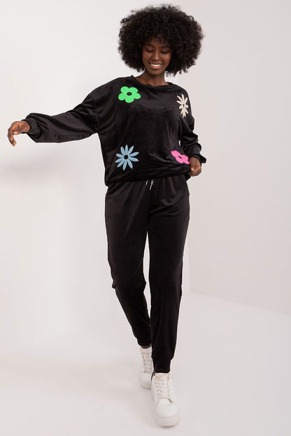 Model presenting the sophisticated black floral velour set by Italy Moda, featuring a plush long-sleeve sweatshirt with colourful floral appliqués, complemented by sleek black high-waisted pants and trendy white sneakers, exemplifying luxury loungewear.