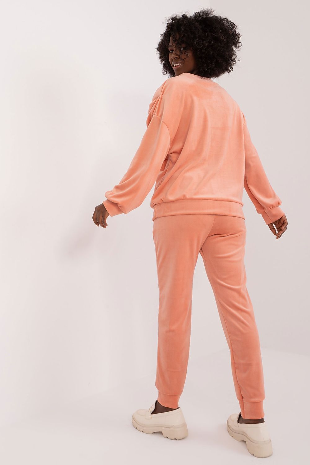 Rear view of Italy Moda velour sweatshirt and pants set in peach colour, showcasing the flowing design and relaxed fit. Model smiles, radiating a chic and fashionable aura.