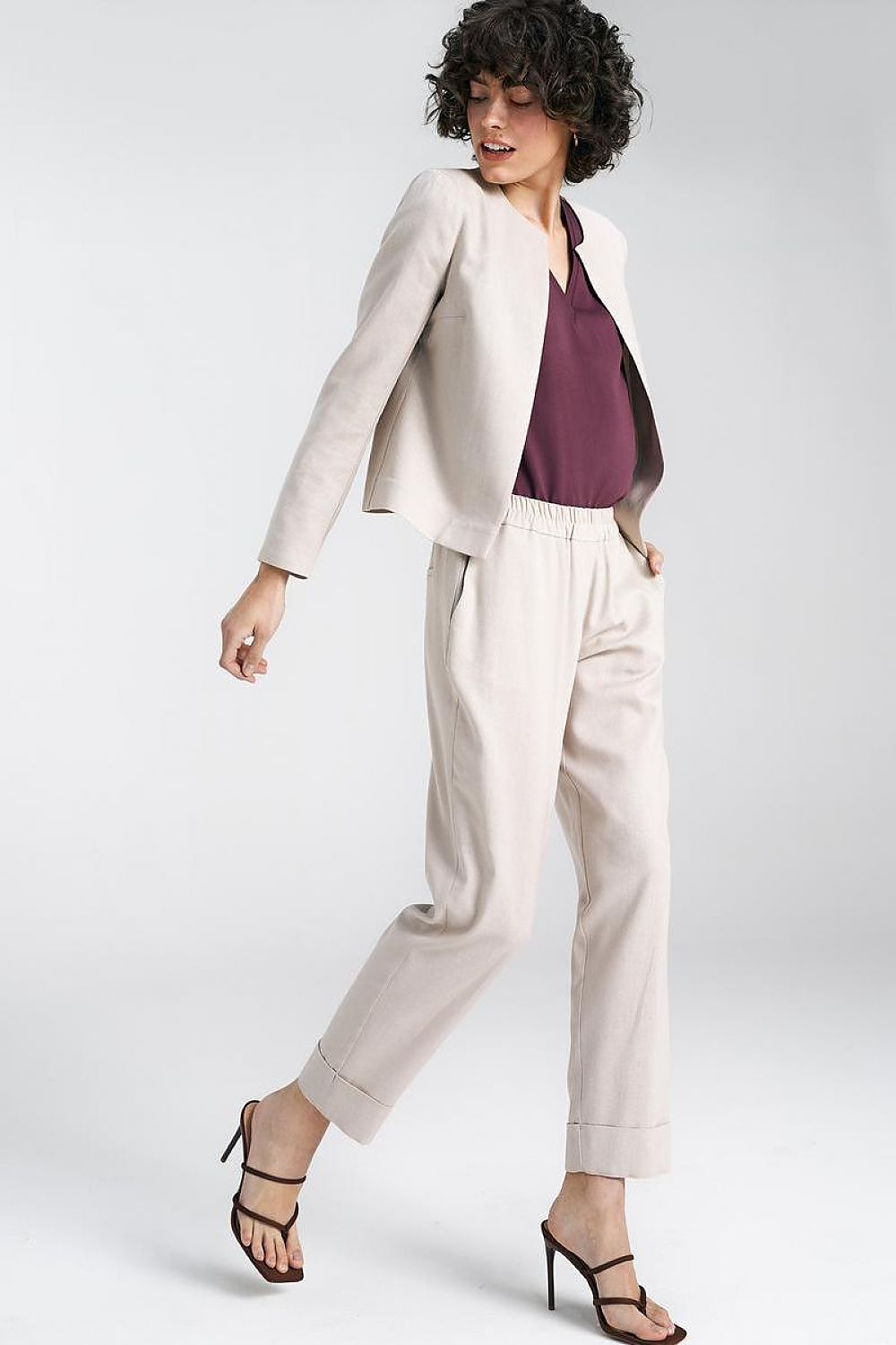 Dynamic pose of a model in a beige jacket and tailored trousers with a maroon top, representing the contemporary style of Nife.