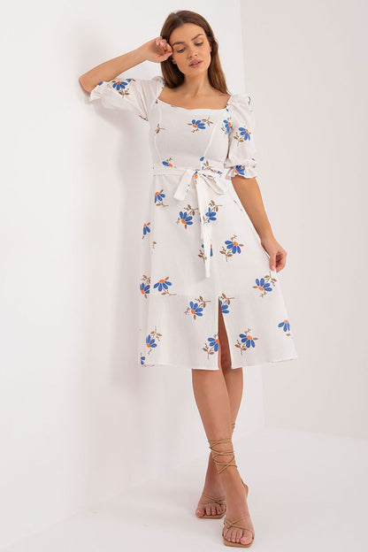 Chic white floral midi summer dress by Lakerta, showcasing a square neckline and a tied waist. The model stands confidently, exuding elegance with blue floral prints, complemented by stylish strappy heels.