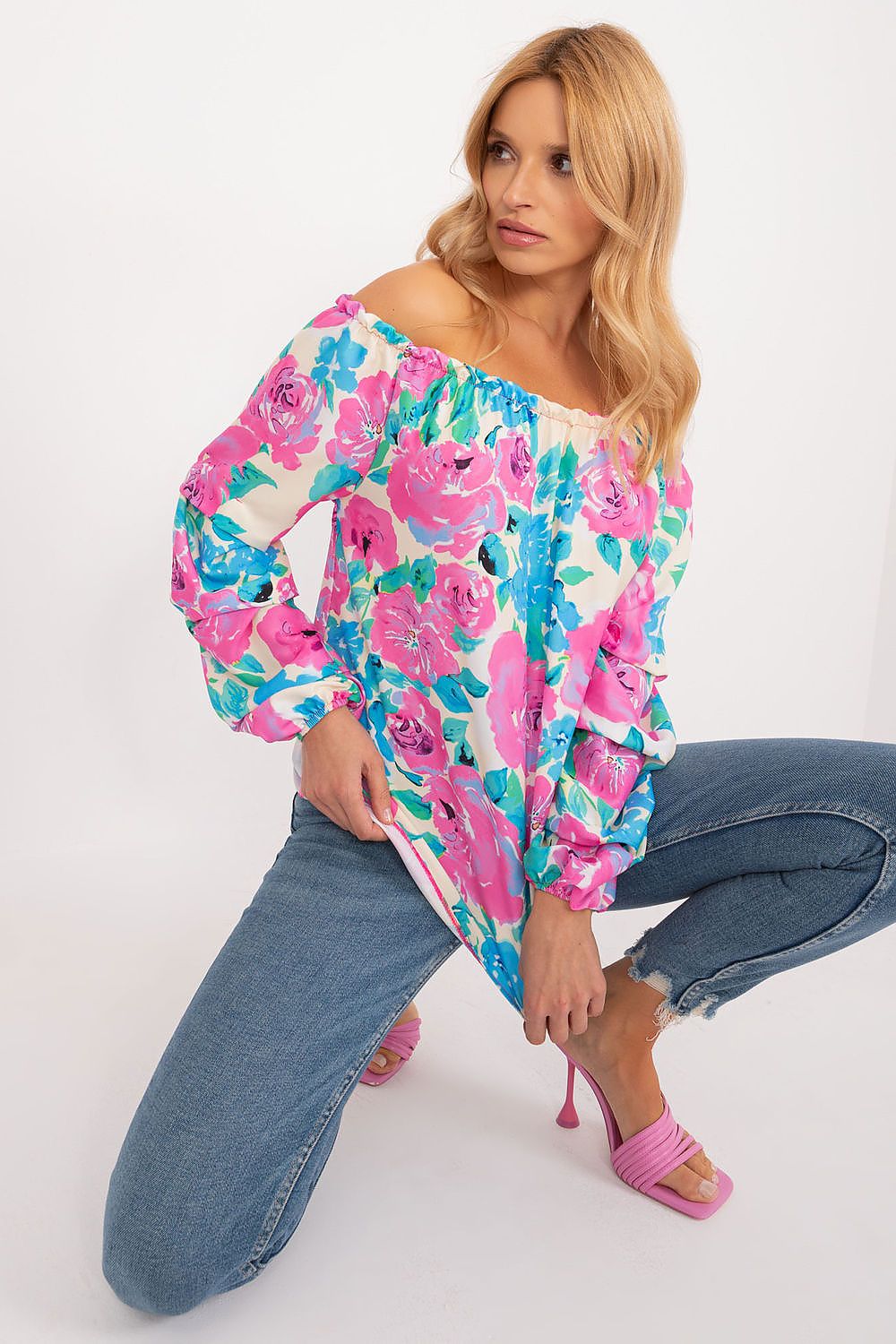 Stylish model wearing a floral print blouse by Rue Paris with off-the-shoulder design, featuring vibrant pink roses against a white background, paired with casual blue jeans and elegant pink heels.