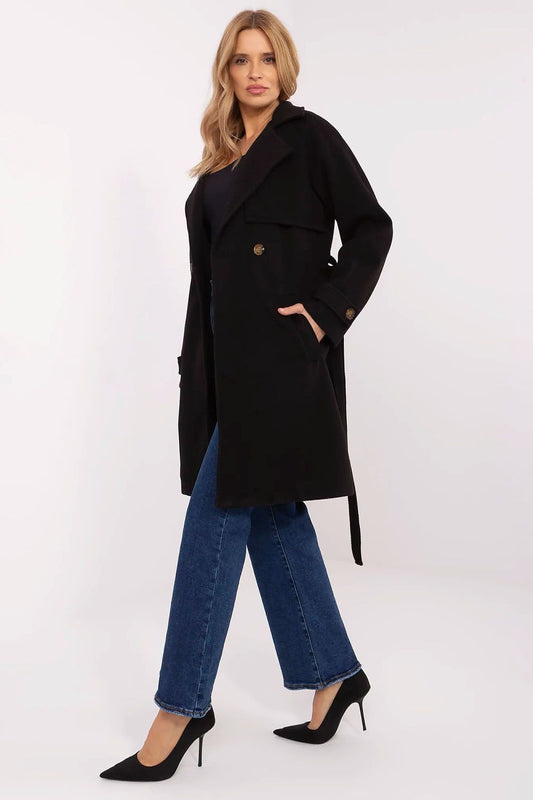 Elegant women's knee-length double-breasted coat in black by Och Bella, featuring a waist tie, styled with denim and heels.
