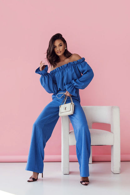 Elegant women's blue ensemble featuring a frilled off-shoulder blouse and high-waisted trousers, designed by LaBalancia, against a soft pink background.