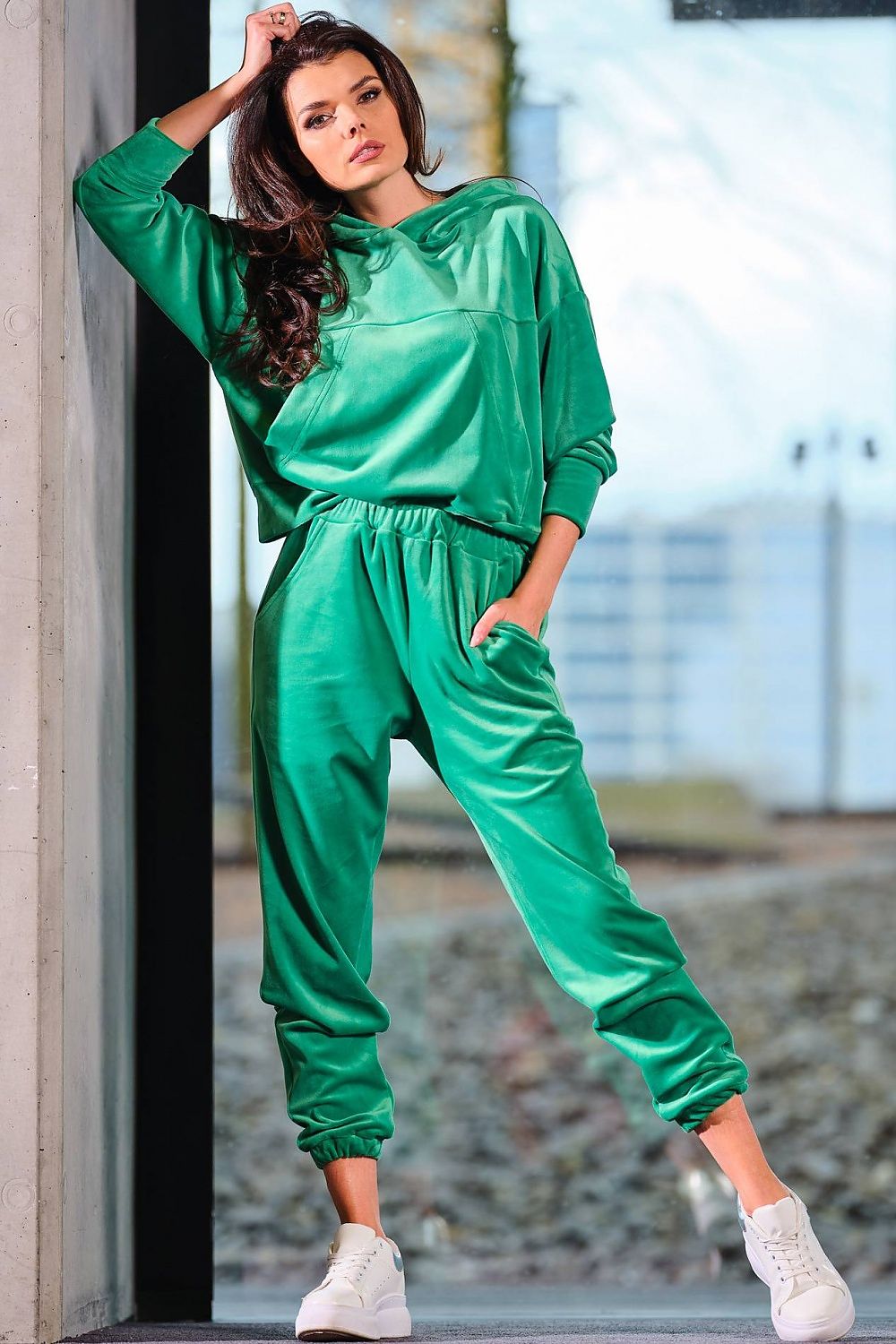 Full-body view of a model in a luxury green velour hooded kangaroo sweatshirt by Awama, captured in a modern outdoor setting, revealing the stylish sportswear look.