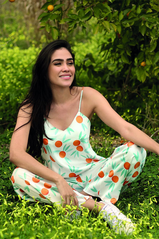 Elegant DKaren satin two-piece pyjamas set featuring a model seated in a lush green environment, adorned with a charming orange and leaf print. The outfit showcases adjustable shoulder straps and a comfortable fit, ideal for luxurious leisure wear.