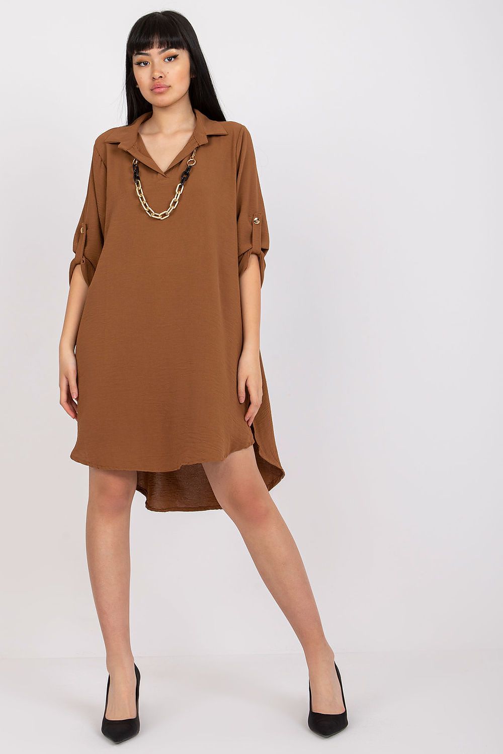 Model showcasing the Asymmetrical Long Sleeve Dress with Collar by Italy Moda in rich brown, prominently featuring a stylish collar, a unique asymmetrical hem, and optional folded sleeves. Enhanced with a chic gold chain accessory, the dress is paired with elegant black high heels against a neutral backdrop.
