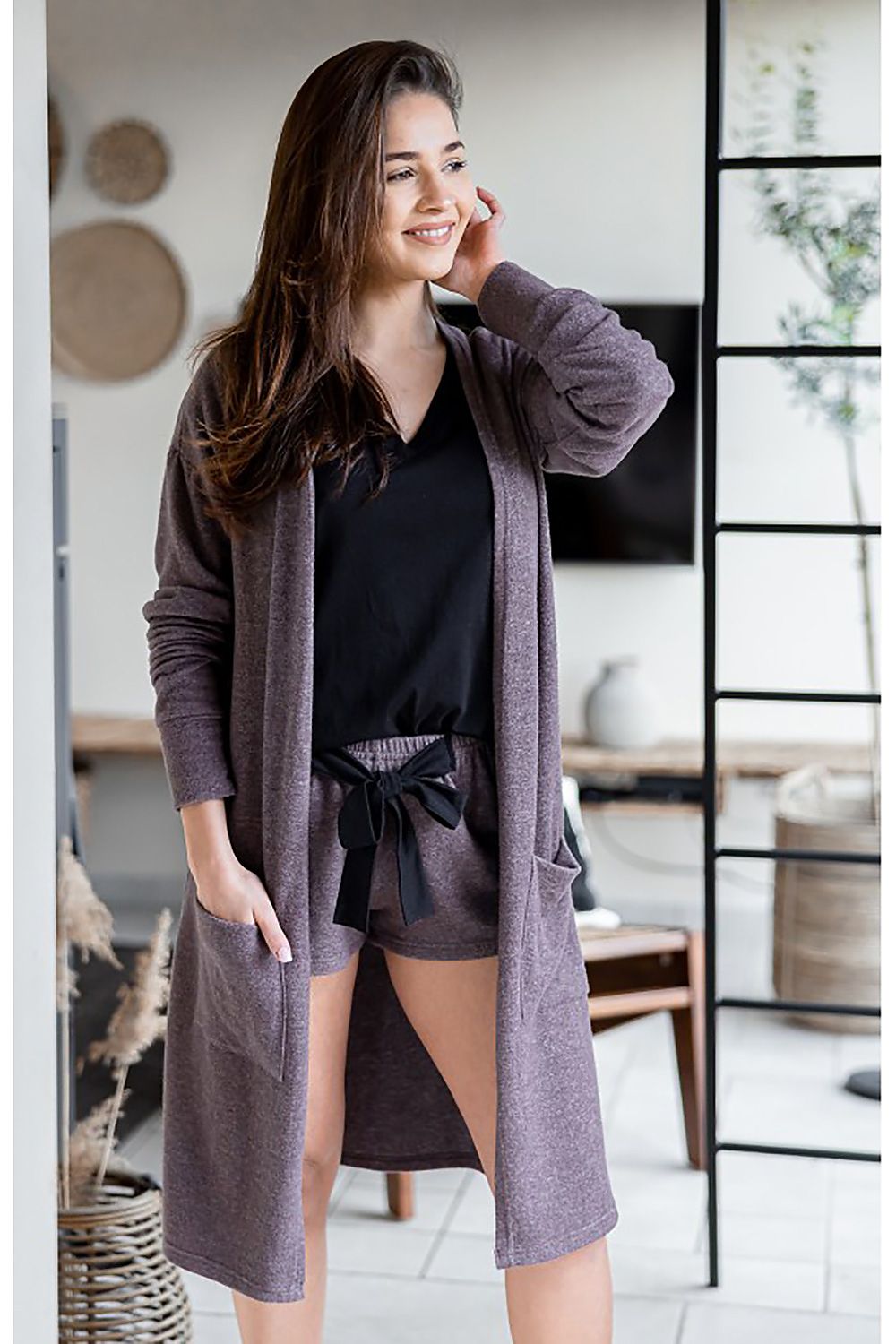 Elegant woman wearing a luxurious soft knit long cardigan by Sensis, showcasing a chic and comfortable style with side pockets, paired with stylish shorts. Ideal for a sophisticated yet casual look in a modern interior setting.