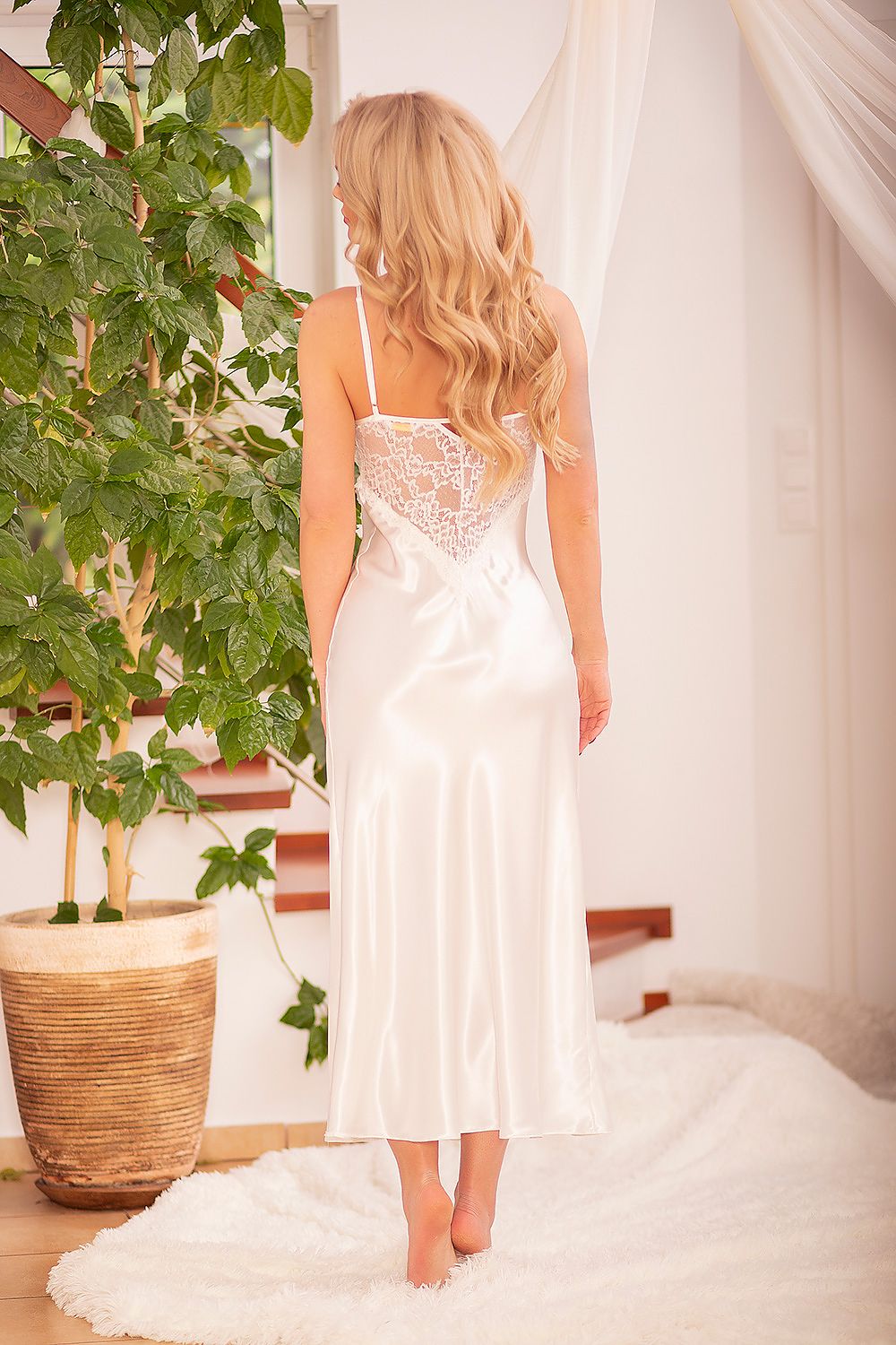 Elegant back view of a beautiful ivory satin slip dress with lace detailing, portrayed in a serene indoor setting surrounded by lush foliage.
