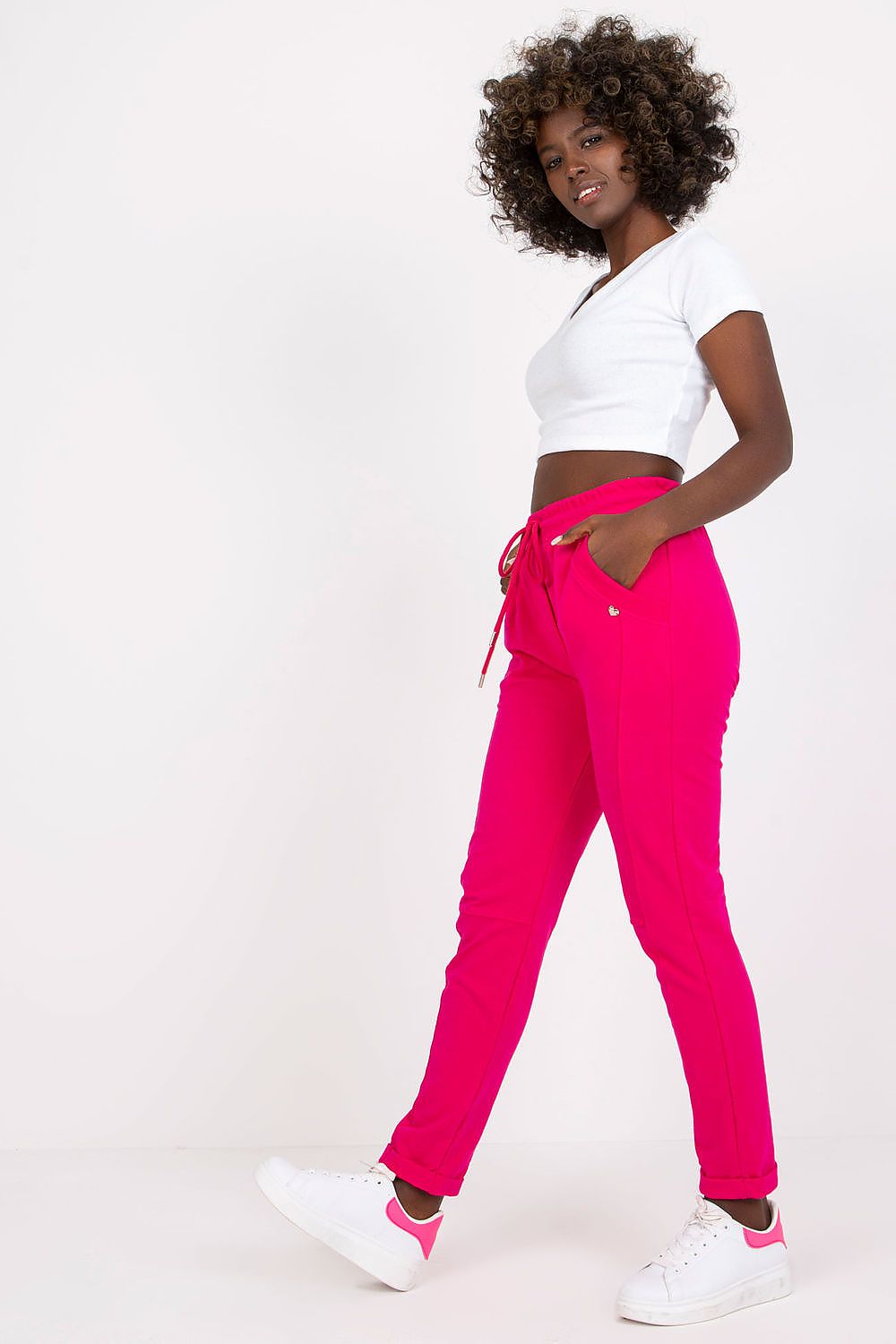 Stylish woman showcasing luxury high-waist BFG tracksuit trousers in vibrant pink, complemented by a fitted white crop top and white sneakers, exuding chic comfort.