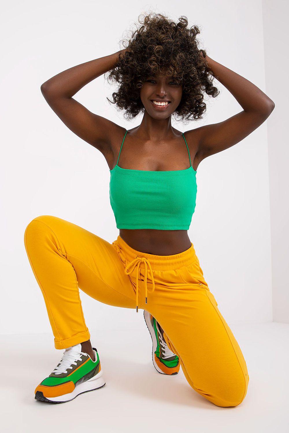 Chic young woman in yellow high-waist tracksuit trousers and a green crop top, striking a dynamic pose, exemplifying modern athleisure style.