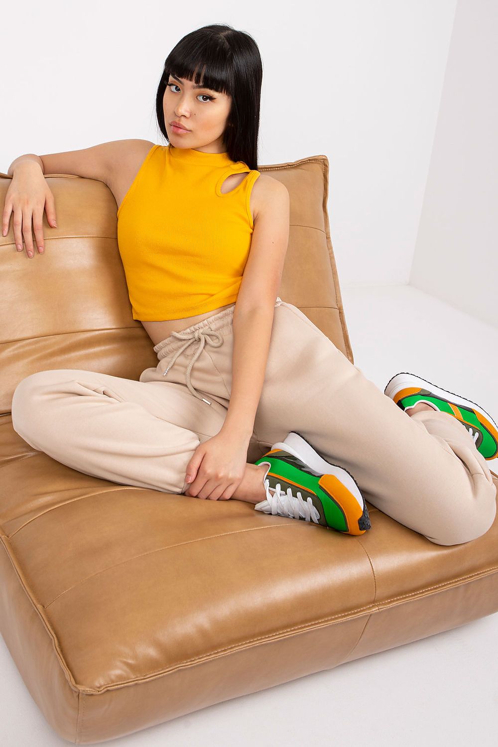 Sophisticated display of Ex Moda Women's Tracksuit Trousers in beige, model seated gracefully on a stylish brown couch wearing a chic yellow crop top and fashionable sneakers, exuding luxury and comfort.