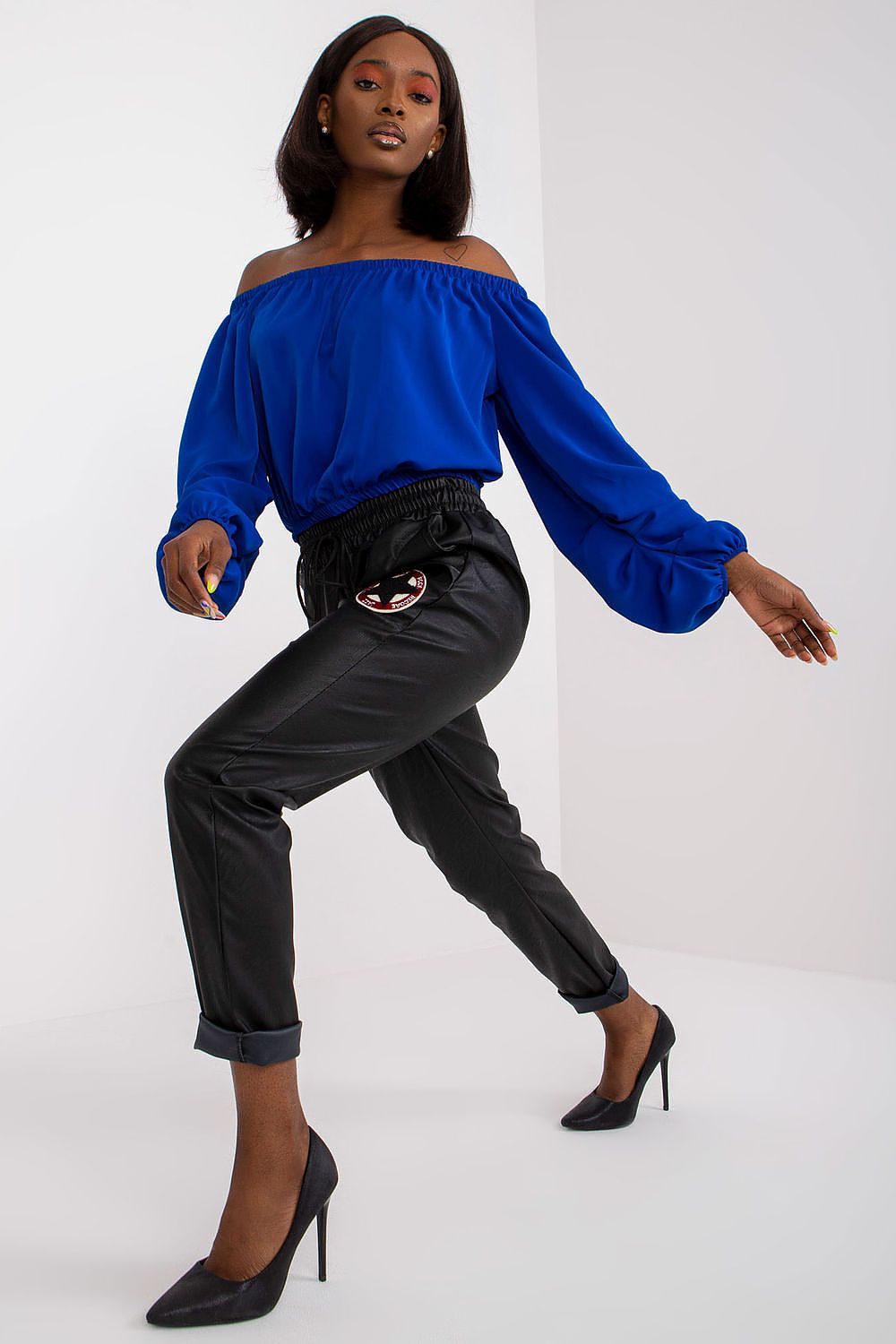 Fashionable model in motion wearing Luxury Women's Ecological Leather Trousers with Elastic Waistband from Italy Moda, paired with a vibrant blue off-shoulder top.