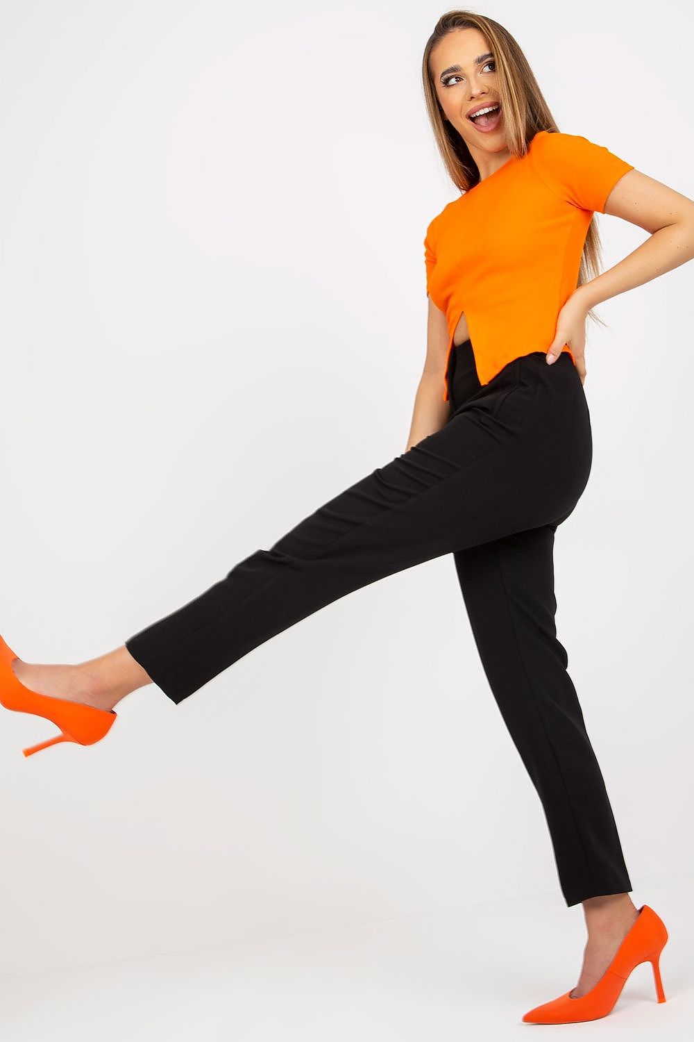A lively pose of a woman in Xsapienza Women’s High-Waisted Straight Cut Trousers, complemented by an orange top and high heels, presenting a fashionable and luxurious style.