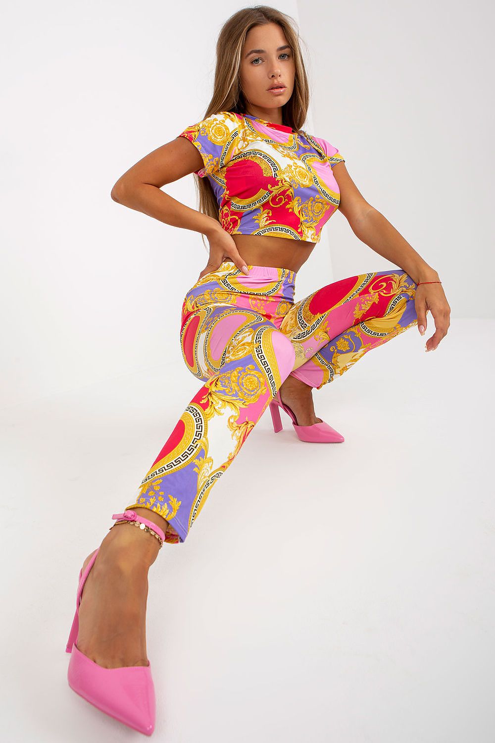 Fashionable woman in luxury high-rise trousers by Italy Moda with an elastic waist, beautifully styled in vivid pink, purple, and gold patterns, striking a dynamic pose in pink heels.