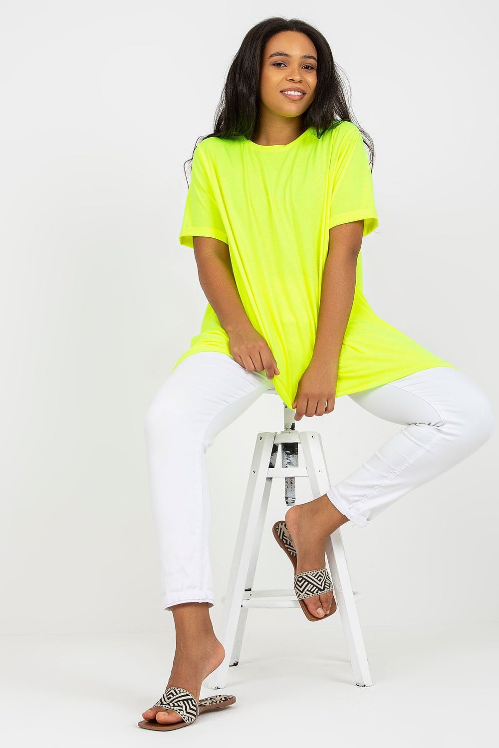 A model showcasing a stylish plus size tunic with short sleeves and an asymmetrical hemline, featuring a boat neck design, paired with white trousers and patterned sandals. Perfect for luxurious everyday styling.