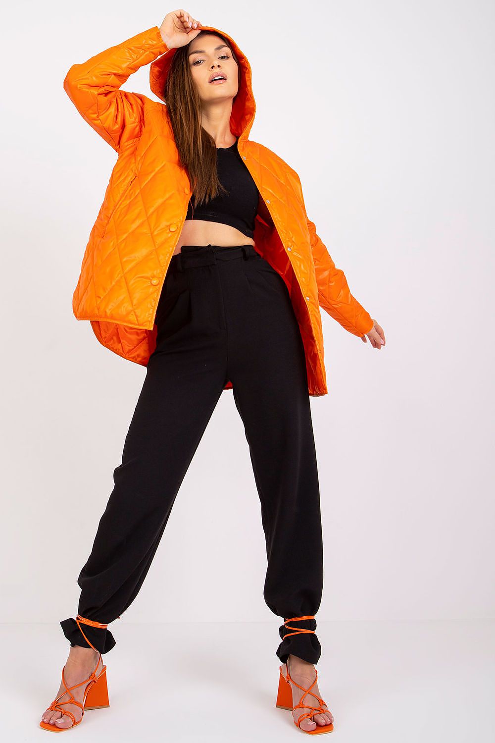 Radiate confidence in the Women's Quilted Hooded Jacket by Rue Paris, in a bold orange hue. The model strikes a fashionable pose, pairing the jacket with chic black trousers, embodying luxury and comfort for transitional seasons.