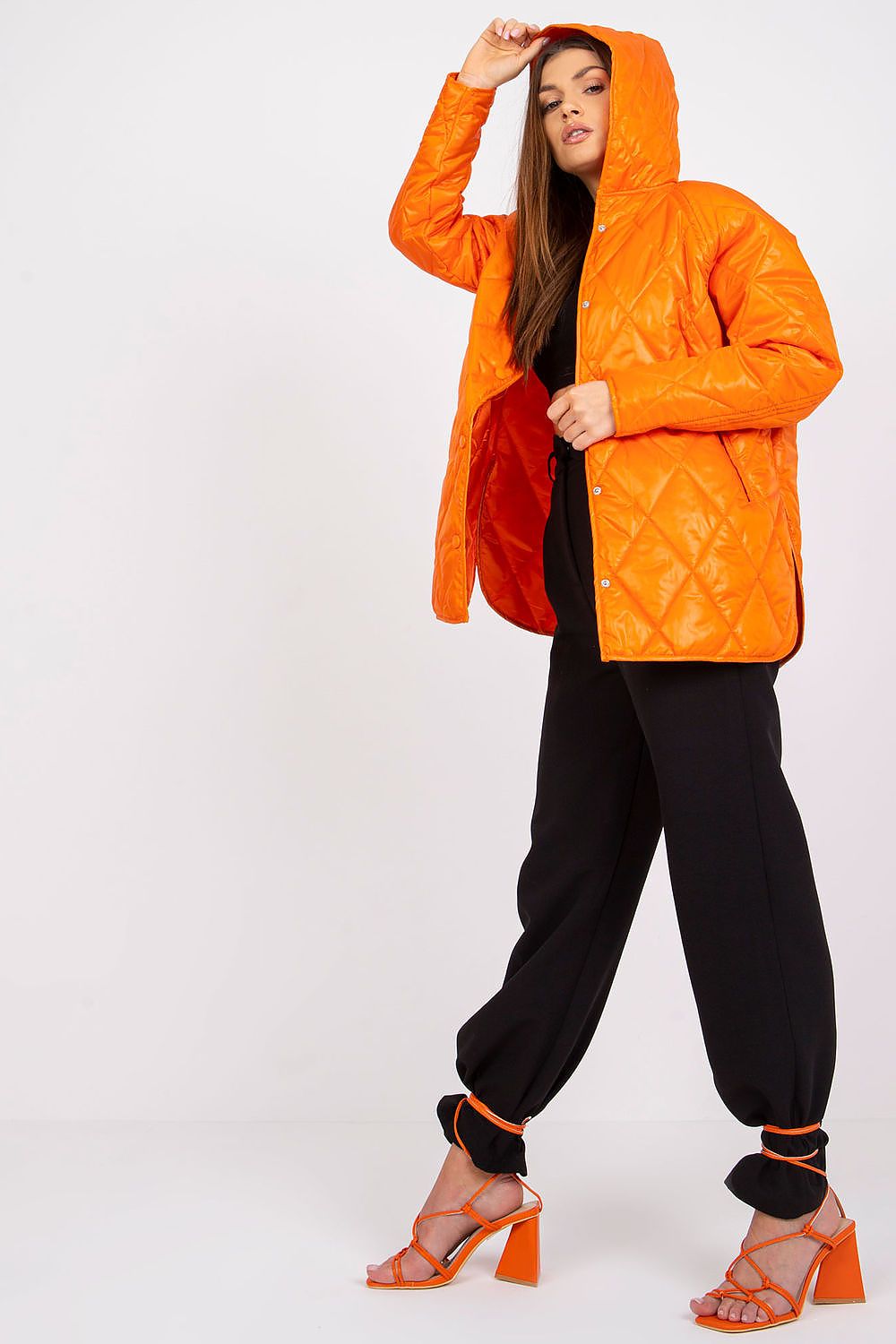 Embrace the trend with the Women's Quilted Hooded Jacket by Rue Paris in vibrant orange. The model confidently showcases the jacket while styled in black trousers, perfect for a sophisticated casual look during transitional weather.