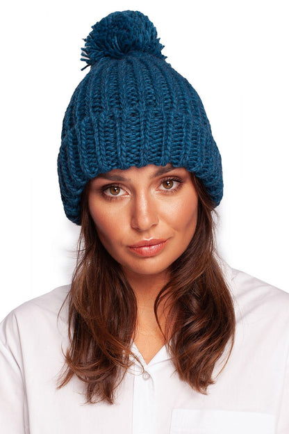 A stunning teal pom-pom beanie hat composed of an exquisite blend of acrylic, nylon, and wool, modelled by a woman, reflecting luxurious winter wear.