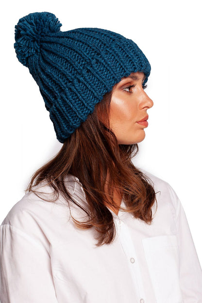 Side perspective of a lavish teal pom-pom beanie hat, elegantly styled and featuring a rich texture, perfect for a fashionable winter look on a model.